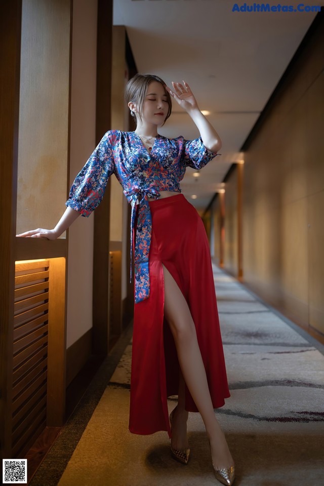 A woman in a blue and red top and red skirt leaning against a wall.